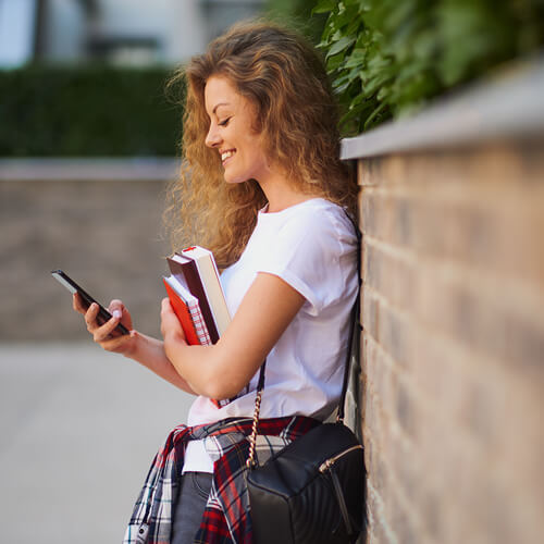 Girl holding books and looking at a smartphone, leaning against a brick wall