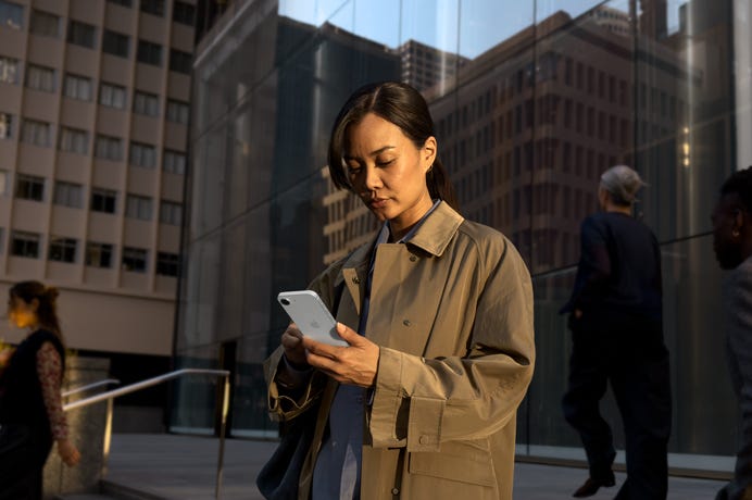 A person using a white iPhone 17e to continue work outside an office building