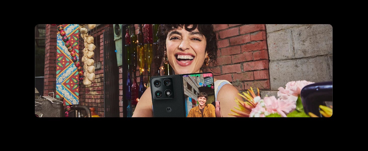 A happy woman holding a partially unfolded Motorola foldable phone showing a smiling man on a video call, against a brick wall decorated with hanging bottles and garlands.