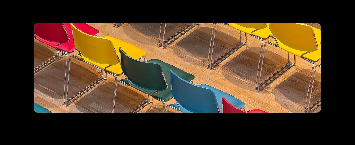 Rows of colourful plastic chairs in red, yellow, green, blue, and pink on a wooden floor, unoccupied and casting shadows.